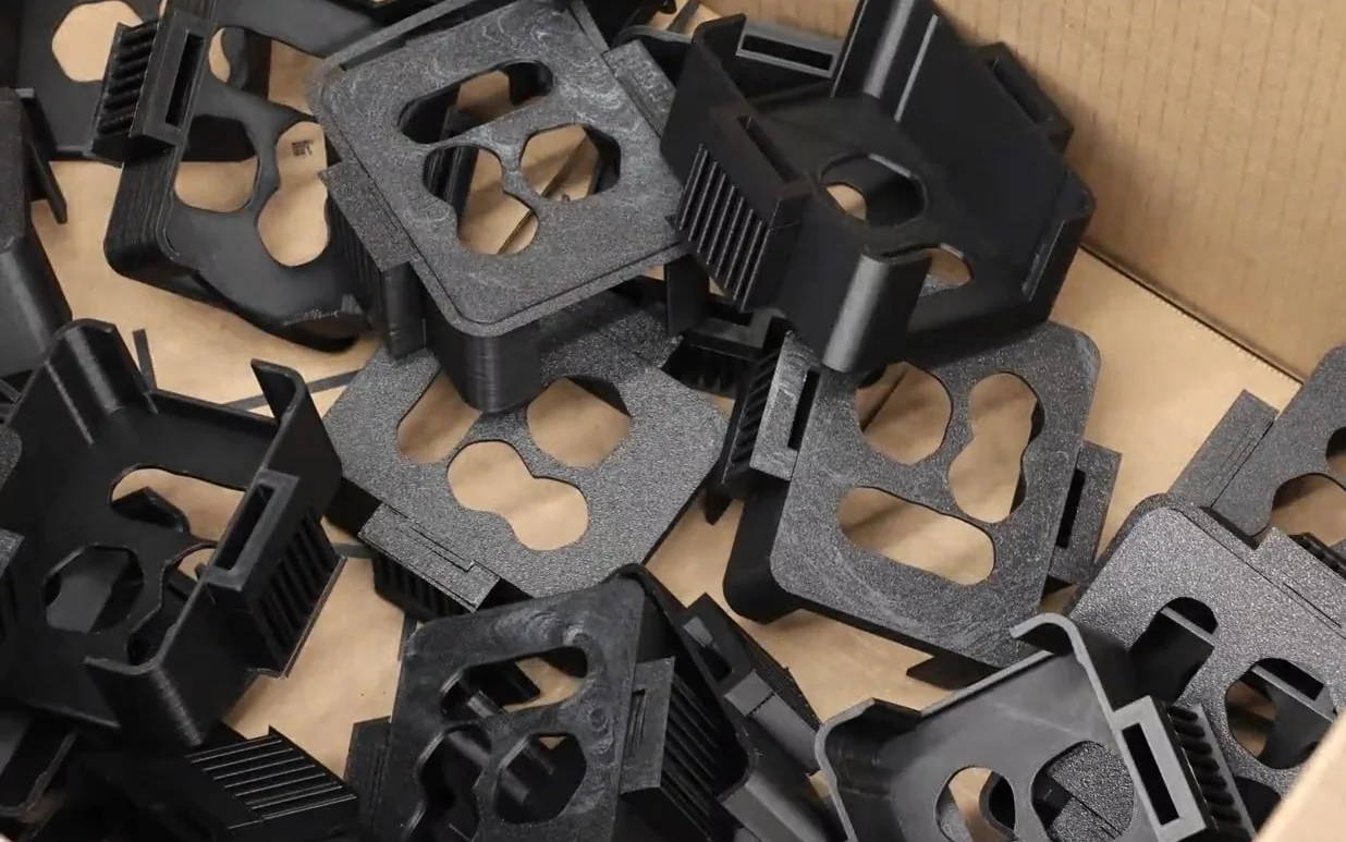 A carton of black 3D-printed brackets fresh off the floor, each with cut-outs and mounting tabs, stacked loose in a shipping box
