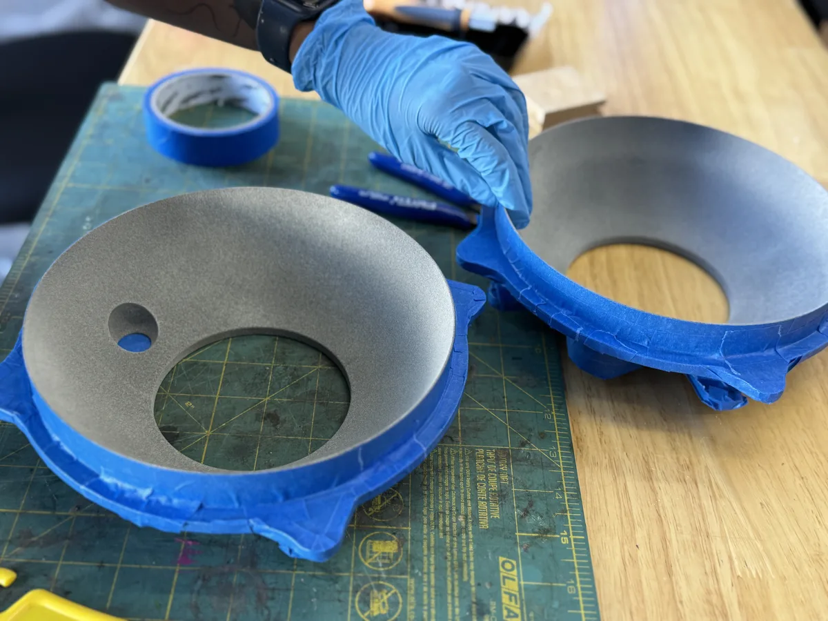 A gloved hand masking two large grey 3D-printed ring-shaped parts with blue painter tape, one on a cutting mat and one on the bench beside it, prepped for painting